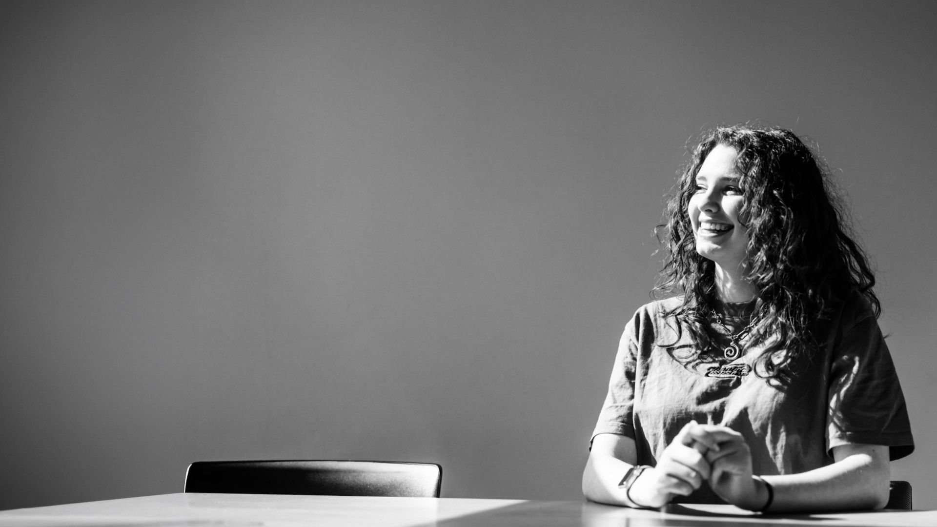 Zwart-witfoto van een jonge vrouw die lachend aan tafel zit, tegen een lege muur